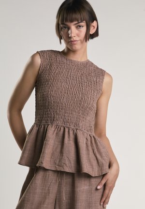 Jonge vrouw met kort bruin haar draagt een mouwloos, gestructureerd bruin peplumtopje en bijpassende broek tegen een effen achtergrond.