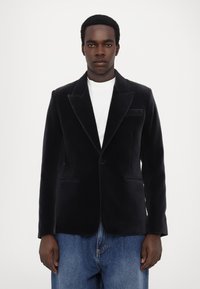 Tamsiai mėlynas aksominis švarkas su įpjovimu, vienu mygtuku ir dviem priekinėmis kišenėmis, derinamas su balta marškinėliais ir džinsiniais kelnais.