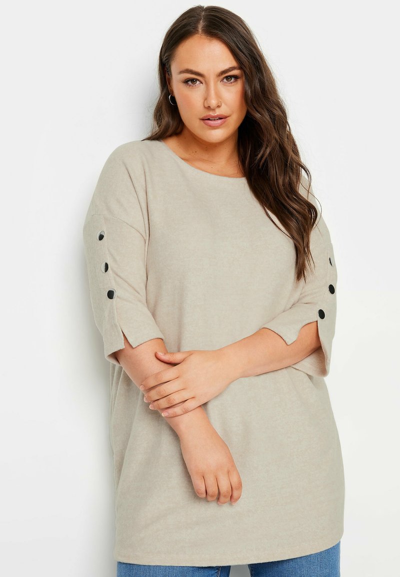 Beige oversized top met een ronde hals, driekwartmouwen en zwarte knoopaccenten, gemaakt van zachte stof met een lichte textuur.