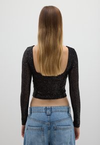 Schwarzes Paillettenoberteil mit langen Ärmeln und tiefem Rückenausschnitt. Der Stoff hat vertikale Details und ist figurbetont, kombiniert mit hoch taillierten Jeans.