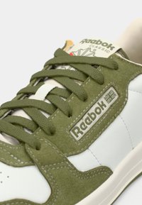 Zelené semišové a bílé kožené tenisky nízkého střihu s kulatou špičkou, plochými šňůrkami, gumovou podrážkou a značkou Reebok na boku.