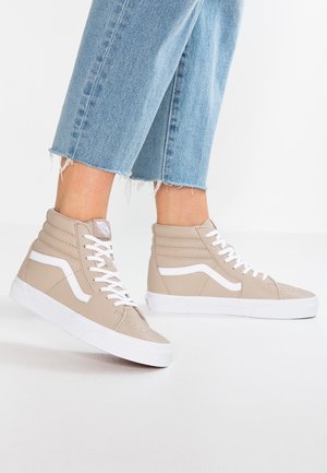 Personne portant des baskets montantes beiges avec des lacets blancs et une bande, associées à un jean bleu raccourci et effiloché sur un sol blanc.