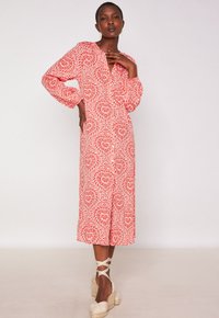 Vrouw in een rood-witte gemusterde midi-jurk met lange mouwen en wedge espadrilles, poserend met één hand op haar heup en de ander bij haar nek.