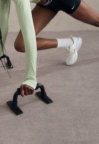 Barre per flessioni nere con dettagli verdi su una superficie beige; una persona che indossa una maglietta a maniche lunghe verde chiaro e pantaloni neri le sta usando.