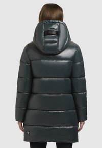 Vrouw in een lange, donkergrijze glanzende pufferjack met een grote capuchon, staande met haar rug naar de camera tegen een effen achtergrond.