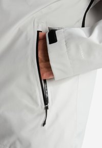 Χέρι μέσα στην τσέπη με φερμουάρ σε λευκό μανίκι μπουφάν με μαύρο ιμάντα Velcro και κείμενο "DRYVENT".