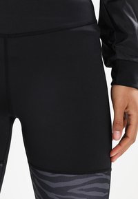 Czarne legginsy sportowe o gładkiej fakturze, z akcentem w postaci czarno-szarego wzoru w paski zebry na dolnej części nogawki. Dopasowany krój.