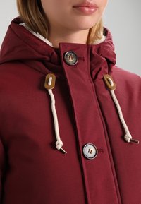 Chaqueta con capucha burdeos forrada de forro polar, con botones de madera, botones metálicos y cordones, que presenta una textura suave.