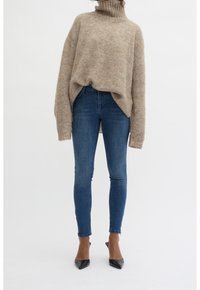 Personne portant un pull beige oversize à col roulé, un jean skinny bleu avec des zips aux chevilles, et des escarpins noirs à bout pointu, se tenant sur un fond blanc.