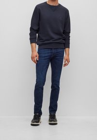 Marinblå tröja med rundad hals och litet bröstlogo, i kombination med mörkblå slim-fit jeans och svarta sneakers med vita detaljer.