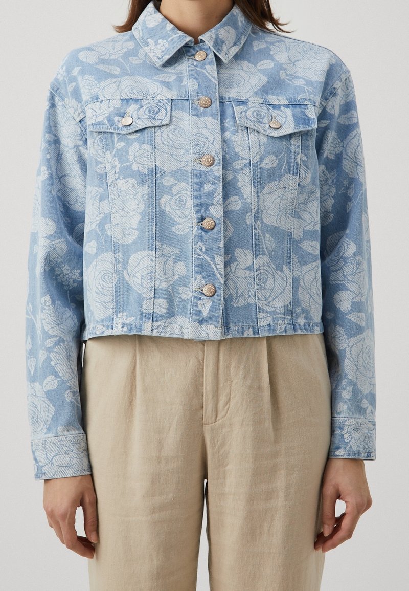 Veste en denim bleu clair avec imprimé floral blanc, boutons métalliques, poches poitrine à rabat, portée avec un pantalon beige plissé taille haute.