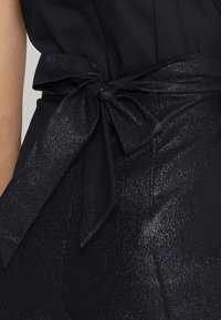 Námořnický overal s lesklým texturovaným páskem uvázaným do mašle v pase, s hladkým tělem z látky.