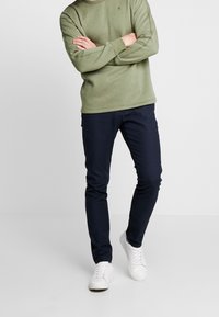 Sudadera verde oliva con cuello redondo, pantalones ajustados azul marino y zapatillas blancas. Tejido suave con detalles de costura sutiles.