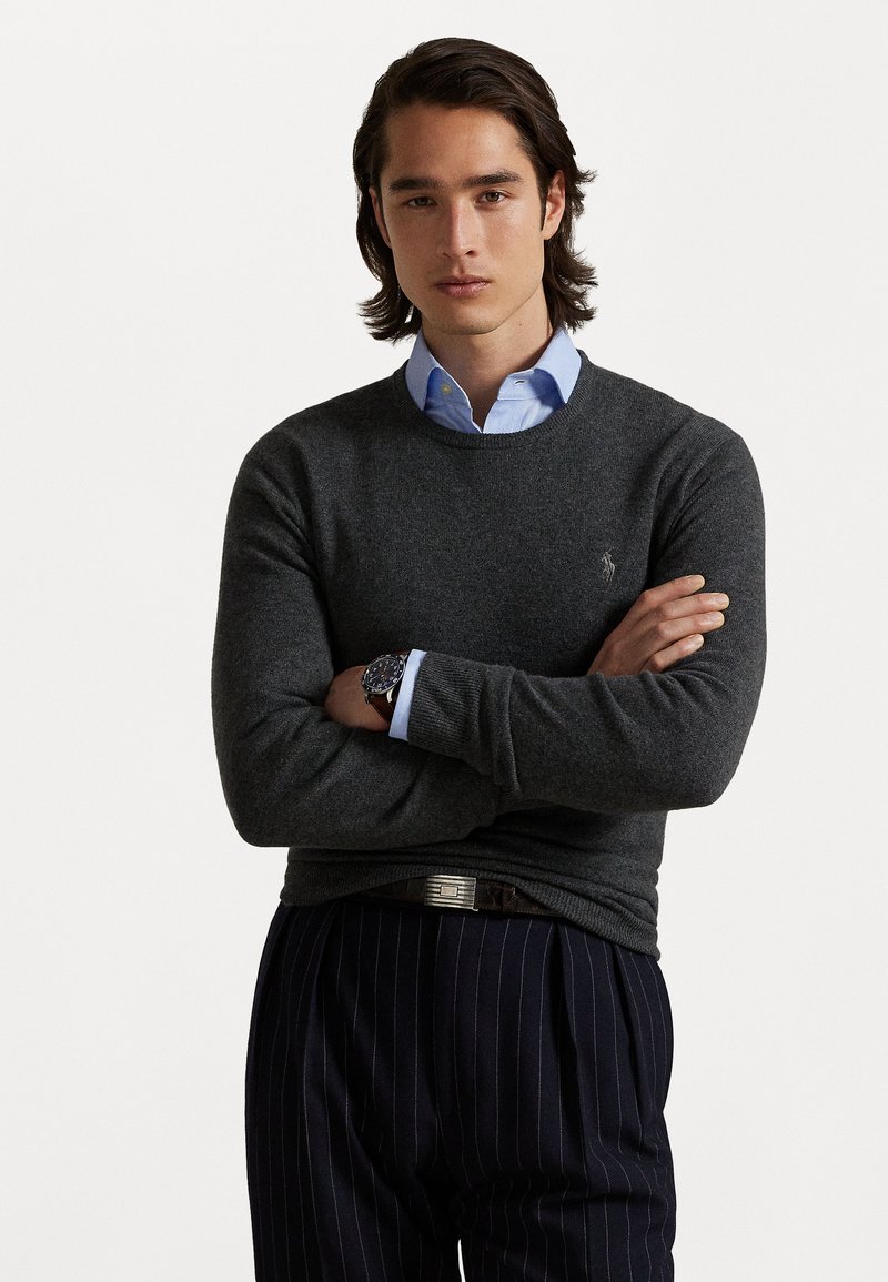 Pull gri închis din tricot cu guler rotund, asociat cu o cămașă cu guler de culoare bleu deschis. Prezintă o croială slim și manșete elasticizate, purtat cu pantaloni cu dungi verticale.
