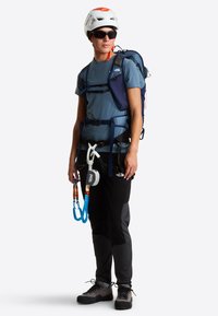 Uprząż wspinaczkowa z pętlami na sprzęt, biały kask, niebieski plecak, szare spodnie i solidne czarne buty. Model stoi wyprostowany w neutralnej pozie.