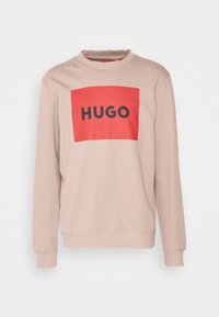 Sudadera beige con cuello redondo y mangas largas, presenta un logotipo rectangular rojo con la palabra "HUGO" en fuente negra en el centro.