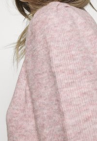 Jasnoróżowy sweter dziergany na drutach, z prążkowaną fakturą, miękkim materiałem i lekko luźnym krojem z marszczeniem przy szwie na ramieniu.