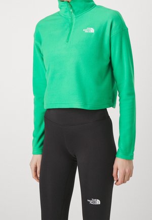 Person trägt eine hellgrüne, kurz geschnittene Fleecejacke mit Viertelreißverschluss und schwarze Leggings, beide mit The North Face Logos.