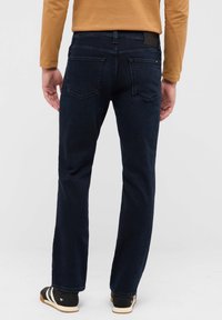 Dunkelblaue Denim-Jeans mit geradem Schnitt. Merkmale sind zwei Gesäßtaschen, dezente Nähte und ein schwarzes Taillenbandetikett. Kombiniert mit dunklen Schuhen.