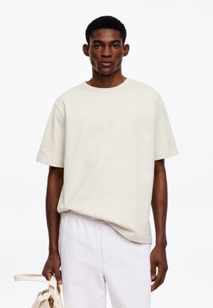 Jeune homme portant un t-shirt beige ample et un pantalon blanc, tenant un sac couleur crème dans sa main gauche sur fond blanc.