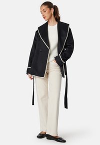 Czarna kurtka z wełny z dużym kołnierzem, białym wykończeniem, podwójnym zapięciem na guziki i związaniami w talii, zestawiona z kremowymi spodniami.