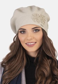 Beret tricotat bej cu vârf adunat, având două aplicații florale și un mic etichetă metalică. Textură netedă, formă rotundă.
