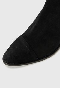 Close-up of a black suede boot, featuring a pointed toe and a smooth surface, with highlighted stitching along the edge.