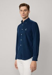 Navyblauwe knoopshirt met een gladde textuur, lange mouwen en een gebogen zoom. Heeft een klein logo op de borst en witte knopen.