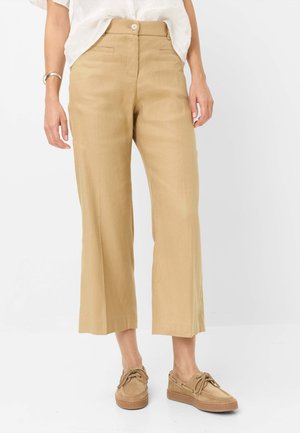 Pantalon beige à jambes larges et coupé, avec bouton et zip devant, porté avec une chemise blanche et des chaussures beiges à lacets sur fond blanc.