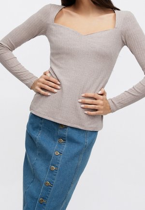 Femme portant un haut beige côtelé à manches longues avec un col carré et une jupe en denim bleu boutonnée, les mains posées sur les hanches.
