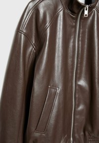 Chaqueta de cuero marrón con cuello alto, cierre de cremallera y un bolsillo lateral. La textura es suave con un ligero brillo.