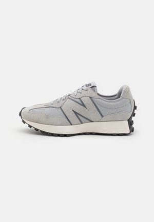Zapatilla New Balance en gris y beige claro con parte superior de ante y malla, cierre de cordones y suela de goma negra texturizada.