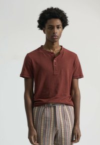 Camisa henley de manga corta de color óxido con un placket de tres botones, combinada con pantalones multicolores estampados con rayas verticales.