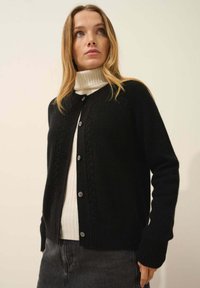 Svart stickad kofta med kabeldetaljer, rund halsringning och knappstängning. Bärs över en vit höghalsskjorta och en grå kjol.