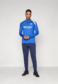 Modrý športový top s dlhým rukávom s bielymi prvkami a logom, v kombinácii s tmavomodrými teplákmi. Outfit dopĺňajú čierne športové topánky.