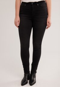 Sieviete valkā pieguļošas melnas džinsa bikses ar augstu jostasvietu un melnus potīšu zābakus, stāvot pret vienkāršu gaišu fonu.