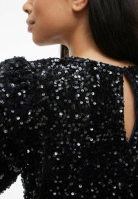 Melns topiņš ar spīdīgu spīguļu apdari un fakturētu virsmu, ar atslēgas caurumu muguras daļā un apjomīgām piedurknēm. Mirdz gaismā.