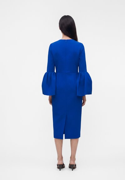 Γυναίκα στραμμένη προς τα πίσω, φοράει εφαρμοστό ρουά φόρεμα με μακριά φουσκωτά μανίκια και σκίσιμο στην πλάτη, συνδυασμένο με μαύρα πέδιλα με τακούνι.