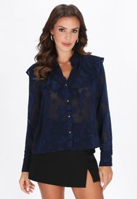 Blusa azul marino transparente con patrón floral, cuello con volantes y cierre de botones. Combinada con una falda negra con una abertura lateral.