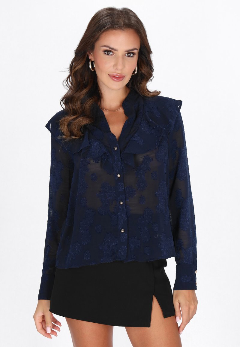Blusa azul marino transparente con patrón floral, cuello con volantes y cierre de botones. Combinada con una falda negra con una abertura lateral.