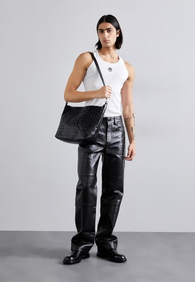 Borsa a spalla nera in pelle con motivi testurizzati, indossata da una persona con una canotta bianca e pantaloni in pelle nera, in piedi contro uno sfondo grigio.
