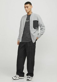 Lys grå zip-jakke med en sort lomme på brystet, parret med en mørkegrå t-shirt og løse sorte cargo-bukser, afsluttet med multicolore sneakers.
