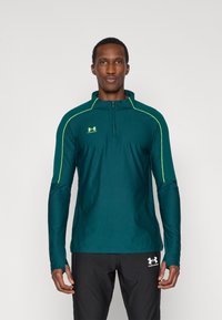 Μακρυμάνικη αθλητική μπλούζα σε τιρκουάζ με γιακά με φερμουάρ μέχρι τη μέση, φωτεινές πράσινες λεπτομέρειες και εφαρμοστή γραμμή. Φοράται με μαύρο αθλητικό παντελόνι.