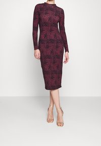 Vestido burdeos de manga larga con patrón floral negro, silueta ajustada, hasta la rodilla. Combinado con tacones dorados con tiras.