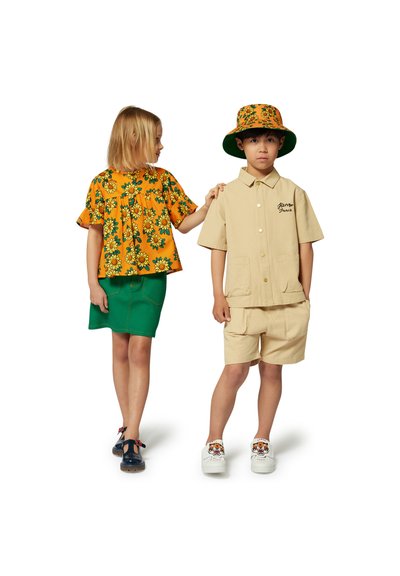 Enfant dans un haut floral jaune avec des manches volantées et une jupe verte ; garçon en chemise beige et short avec logo, portant un chapeau seau assorti.