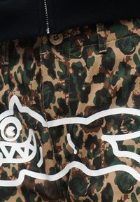 Καμουφλάζ παντελόνι με πράσινο, καφέ και μαύρο μοτίβο, που διαθέτει ένα λευκό γραφικό με το πρόσωπο ενός καρτούν τέρατος και κυματιστή γραμμή σε όλο το ύφασμα.