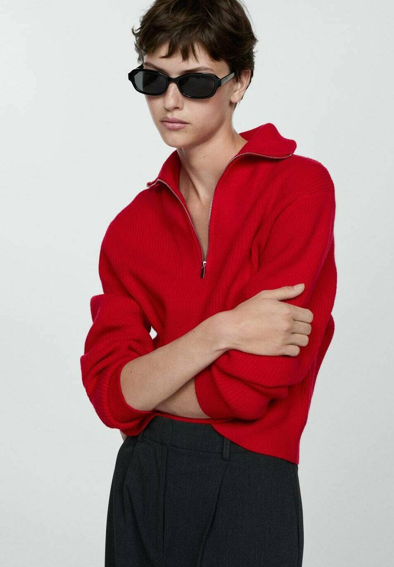 Crvena rebrasta pletena bluza s visokim ovratnikom i patentnim zatvaračem, nosi se preko tamnih hlača. Model nosi crne sunčane naočale, ruke prekrižene.