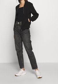 Giacca bomber nera, top nero arricciato, jeans scuri a vita alta e sneaker bianche con suola color gomma. Tessuto liscio e design aderente.