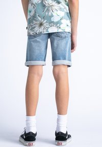 Pantalones cortos de mezclilla azul claro con dobladillos enrollados, vista desde la parte posterior. Llevados con zapatillas Vans negras y calcetines blancos. Camisa floral parcialmente visible.