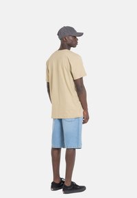 Camiseta de algodón marrón claro y pantalones cortos de denim azul claro, combinados con una gorra gris oscuro y zapatillas negras, vistos desde la parte posterior.